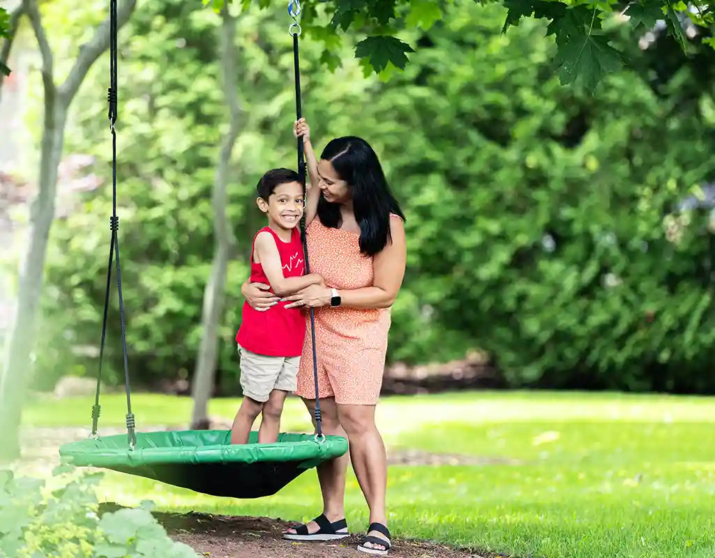 young boy standing on a tree swing with his mother holding him safely