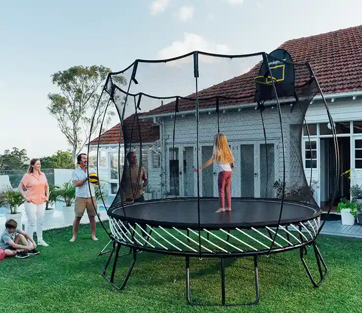 A young girl standing on an outdoor trampoline with her family watching her
